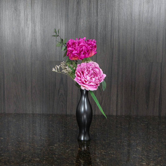 two dried pink peonies, filler, and dried greenery in a black ceramic bud vase