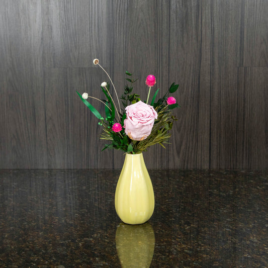 a single light pink preserved rose with dried greenery in a yellow bud vase