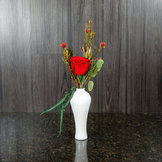 a single red preserved roses and dried greenery in a white bud vase