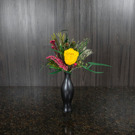a single yellow preserved rose with dried greenery in a black ceramic bud vase