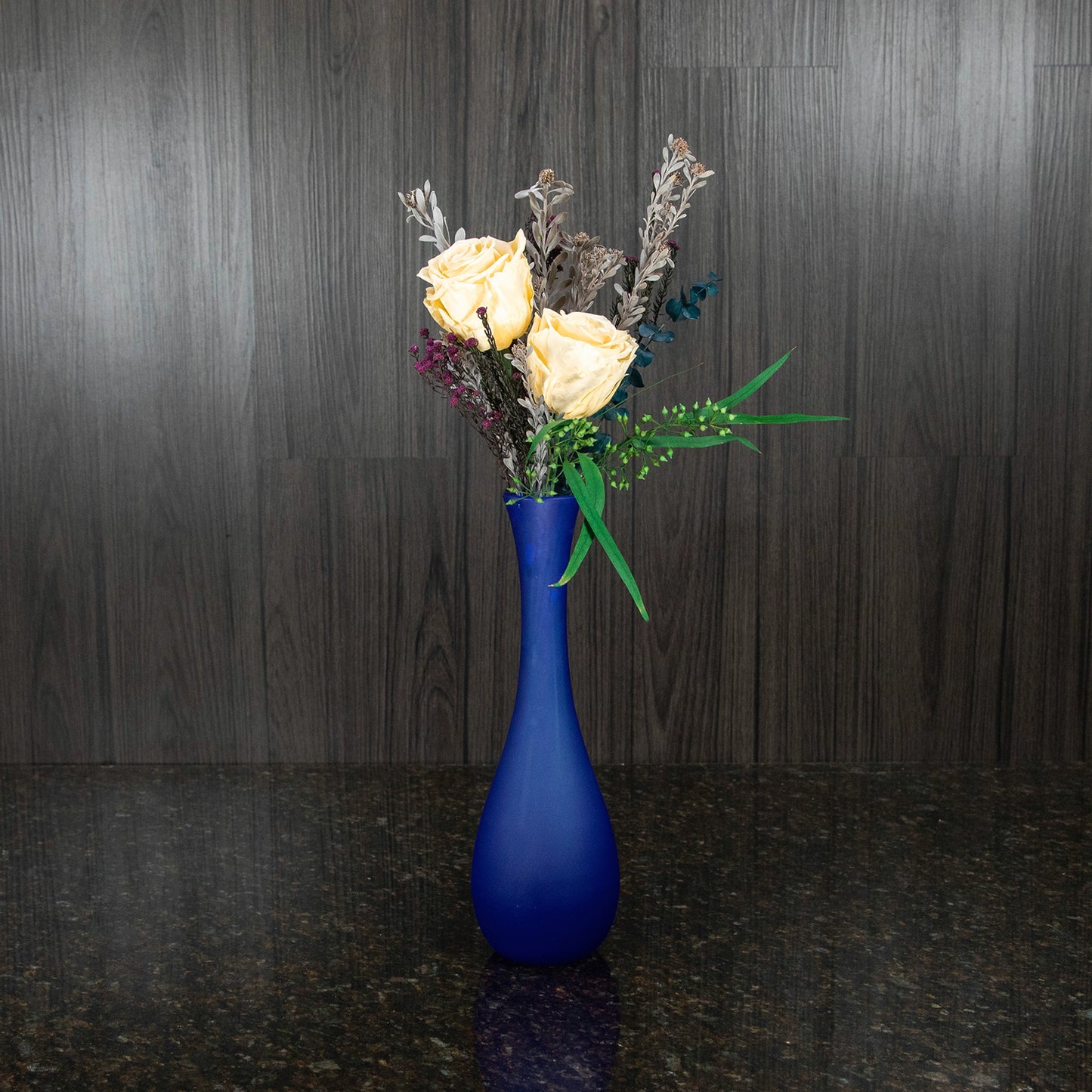 two cream preserved roses with greenery in a tall frosted blue glass vase