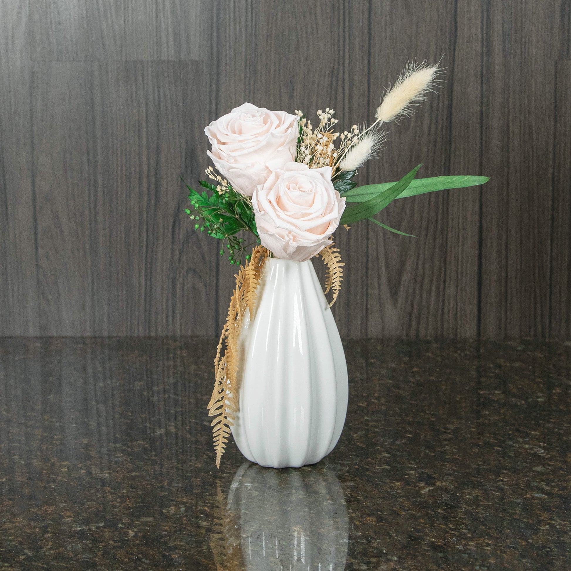 two blush preserved roses with dried, bleached greenery in a white vase