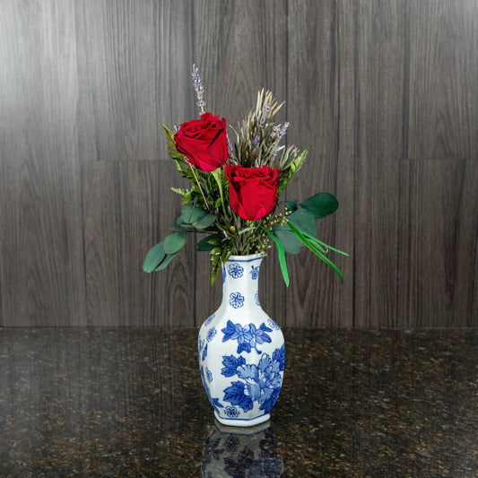 two red preserved roses with dried greenery in a white and blue ceramic vase