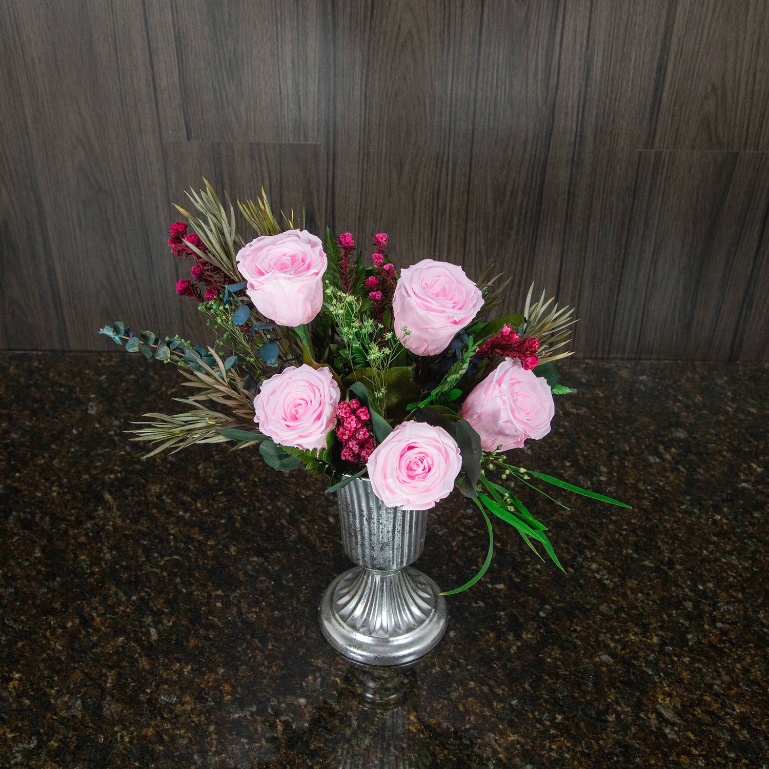 top down view of five pink preserved roses and dried greenery in a footed silver metal vase