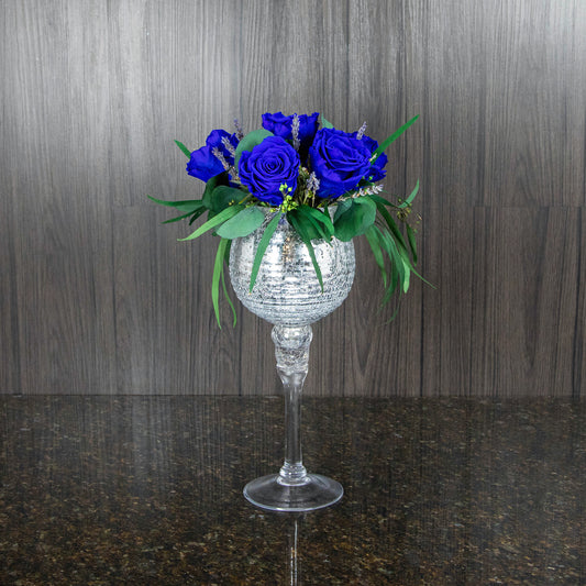 a preserved rose arrangement featuring blue roses and greenery in a tall silver stemmed vase