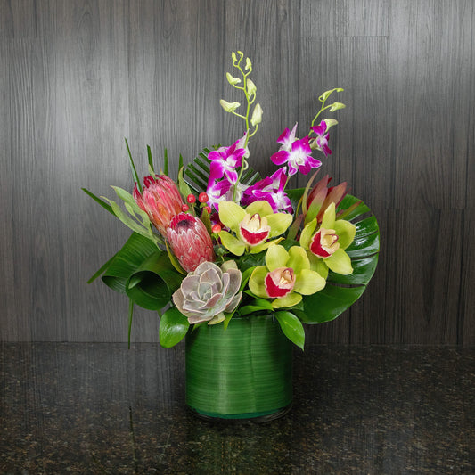 a tropical flower arrangement with a mix of colorful blooms and leaves in a glass cylinder vase