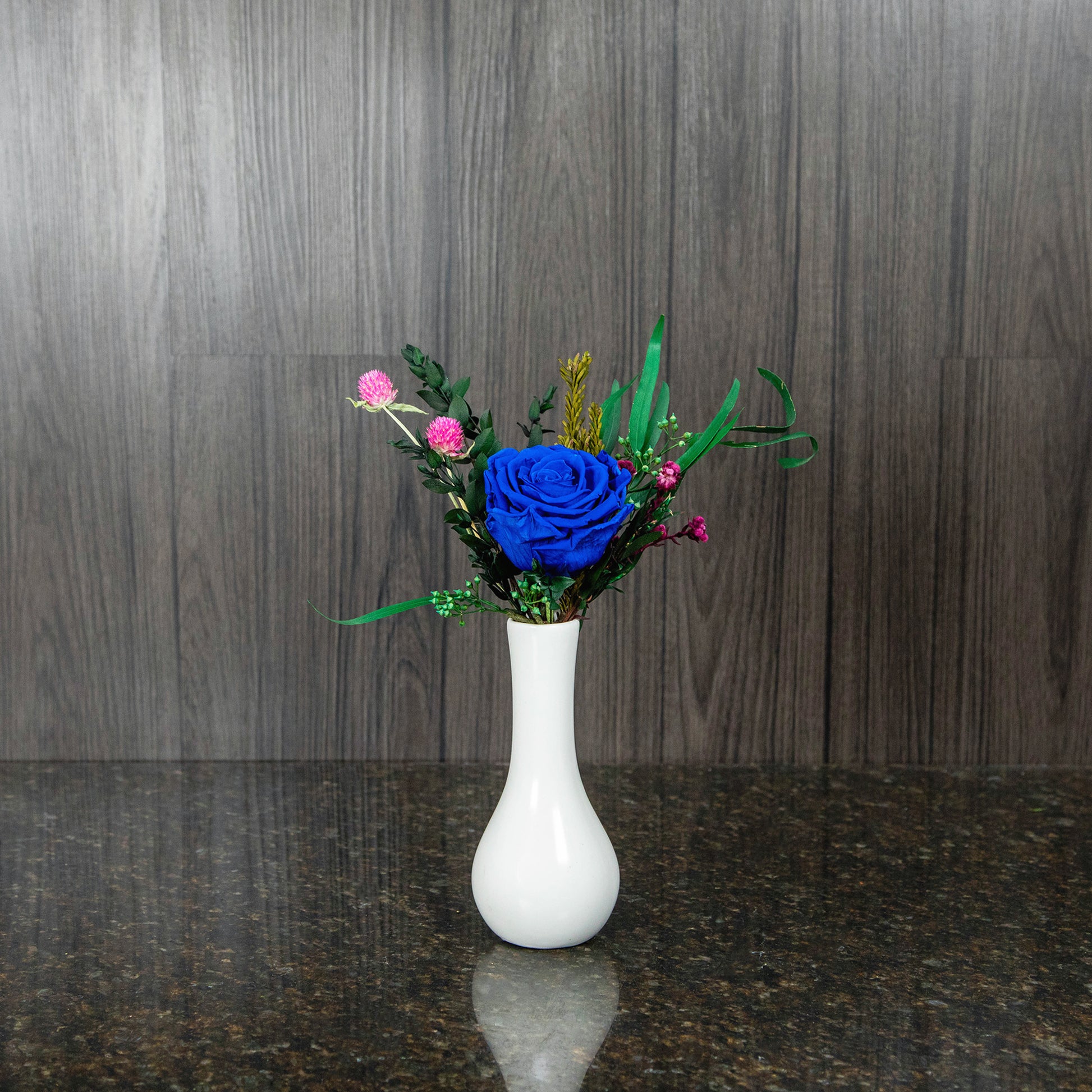 a single blue preserved rose in a white bud vase with bits of preserved greenery and filler