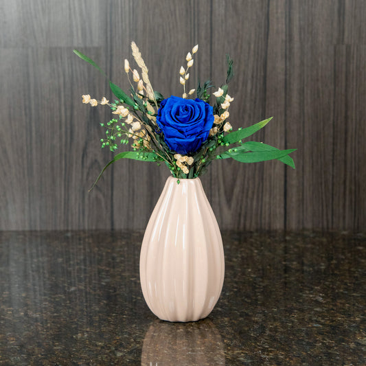 a single blue preserved rose with dried greenery and filler in a beige vase