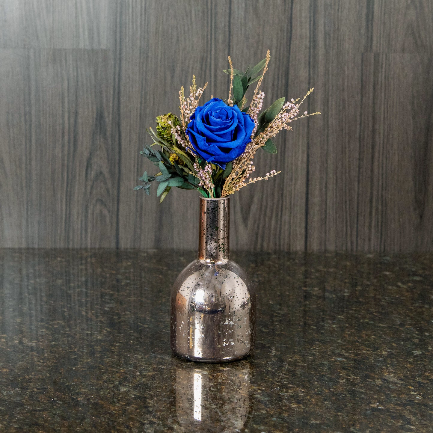 a single blue preserved rose with dried greenery and filler in a rose gold mercury glass bud vase