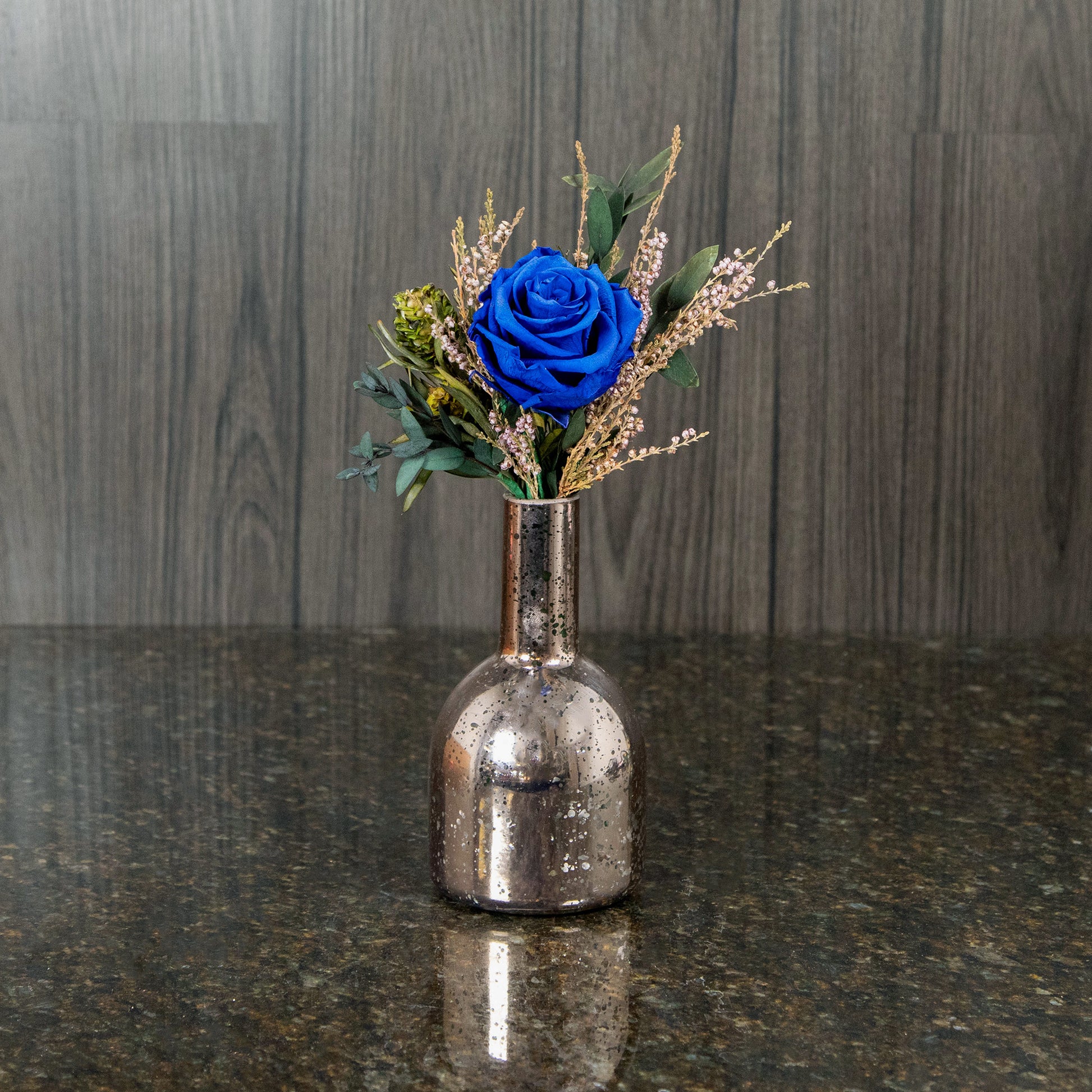 a single blue preserved rose with dried greenery and filler in a rose gold mercury glass bud vase