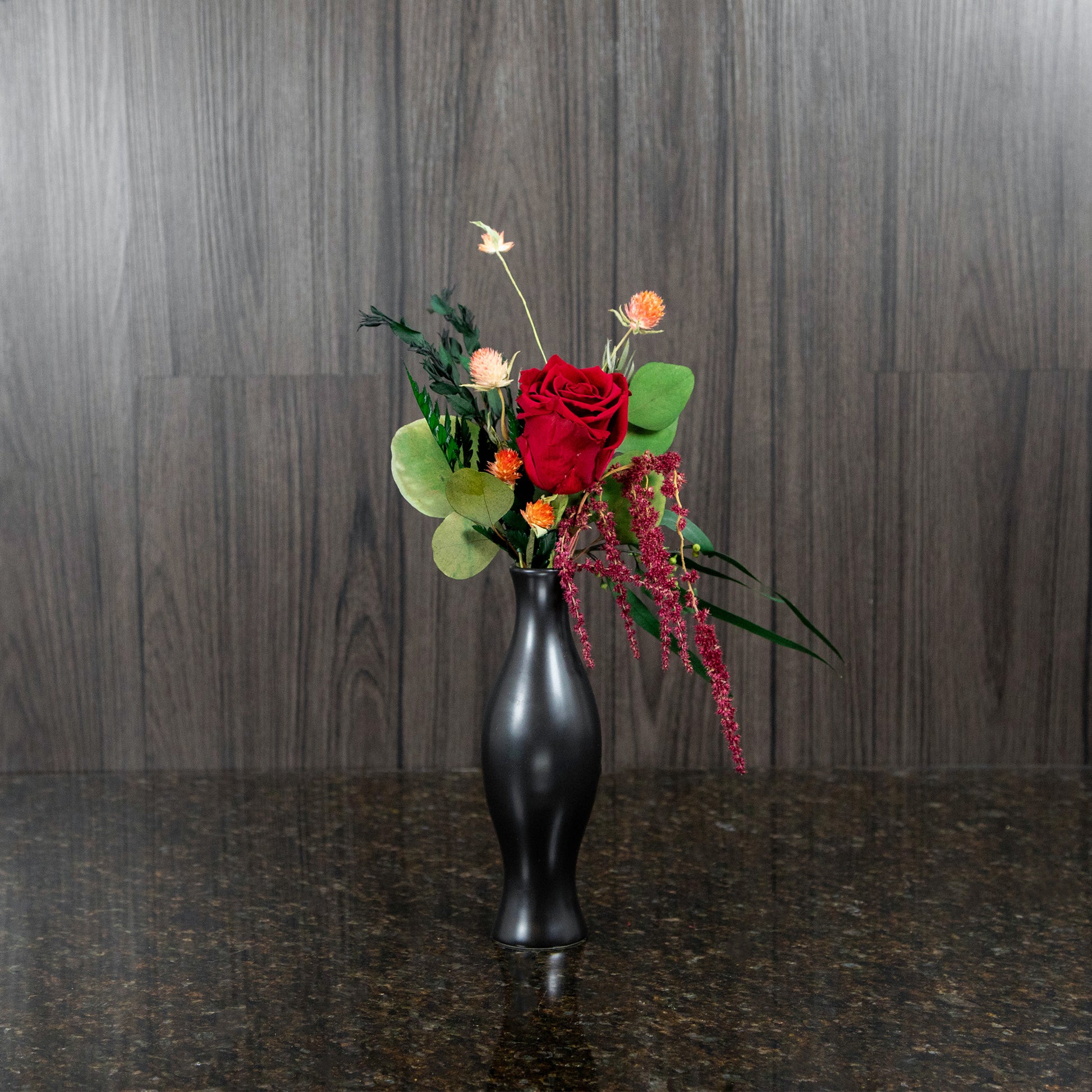 a single red preserved roses and dried greenery in a black bud vase