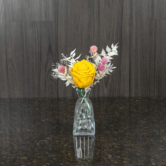 a single yellow preserved rose in a glass bud vase with a bit of preserved greenery and filler