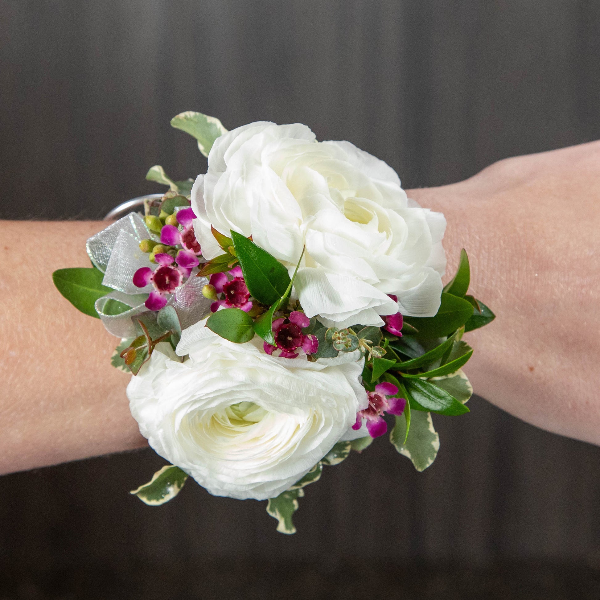 a wrist corsage made with white ranunculus, purple waxflower, and different types of greenery