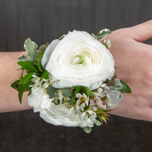 a wrist corsage made with white ranunculus, white waxflower, and different types of greenery