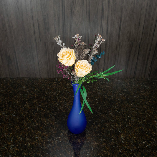 top down view of two cream preserved roses with greenery in a tall frosted blue glass vase