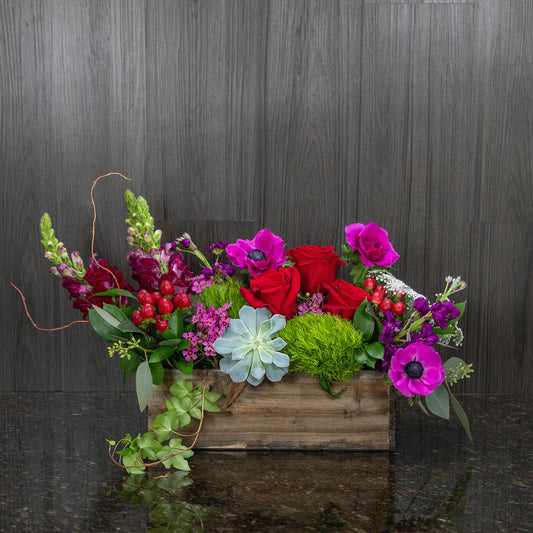 a compact flower arrangement in a wooden box with red, pink, and green flowers and a succulent