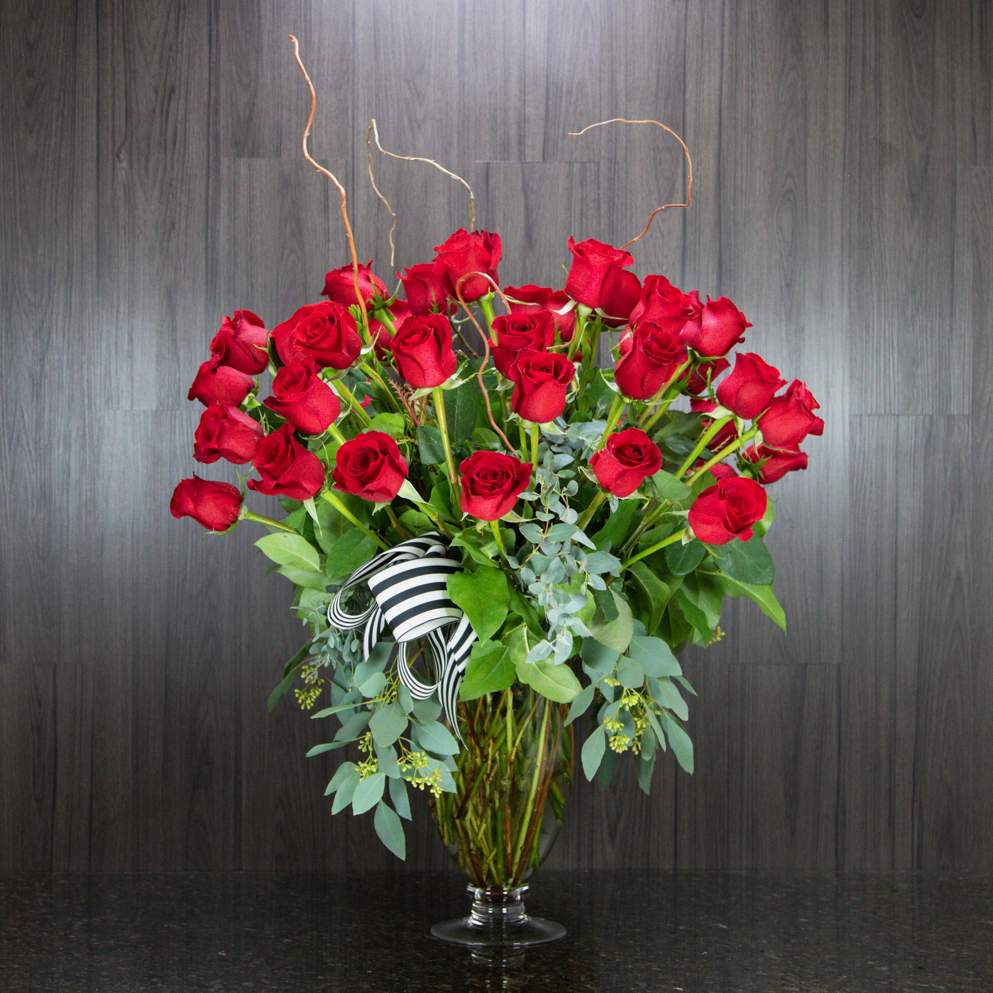 a large display of three dozen red roses with tall stems of curly willow in a glass footed vase 