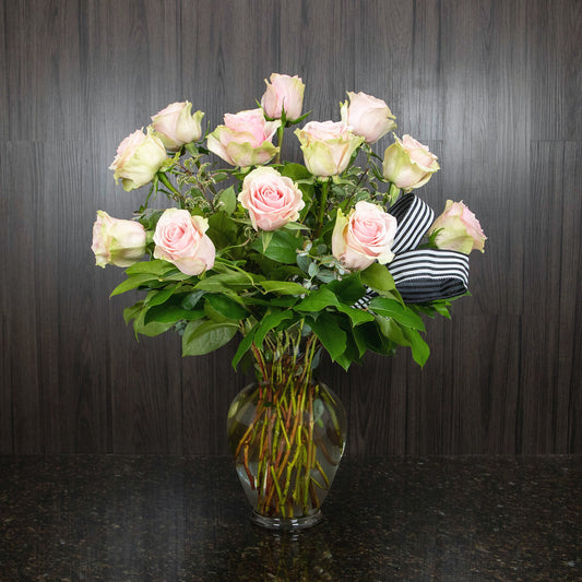 a glass vase with greenery and one dozen light pink roses