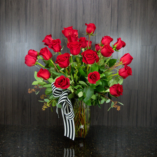 a large display of two dozen red roses with greenery in a glass vase