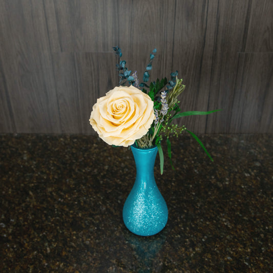 top down view of a single large preserved cream rose and preserved greenery in a blue sparkling vase