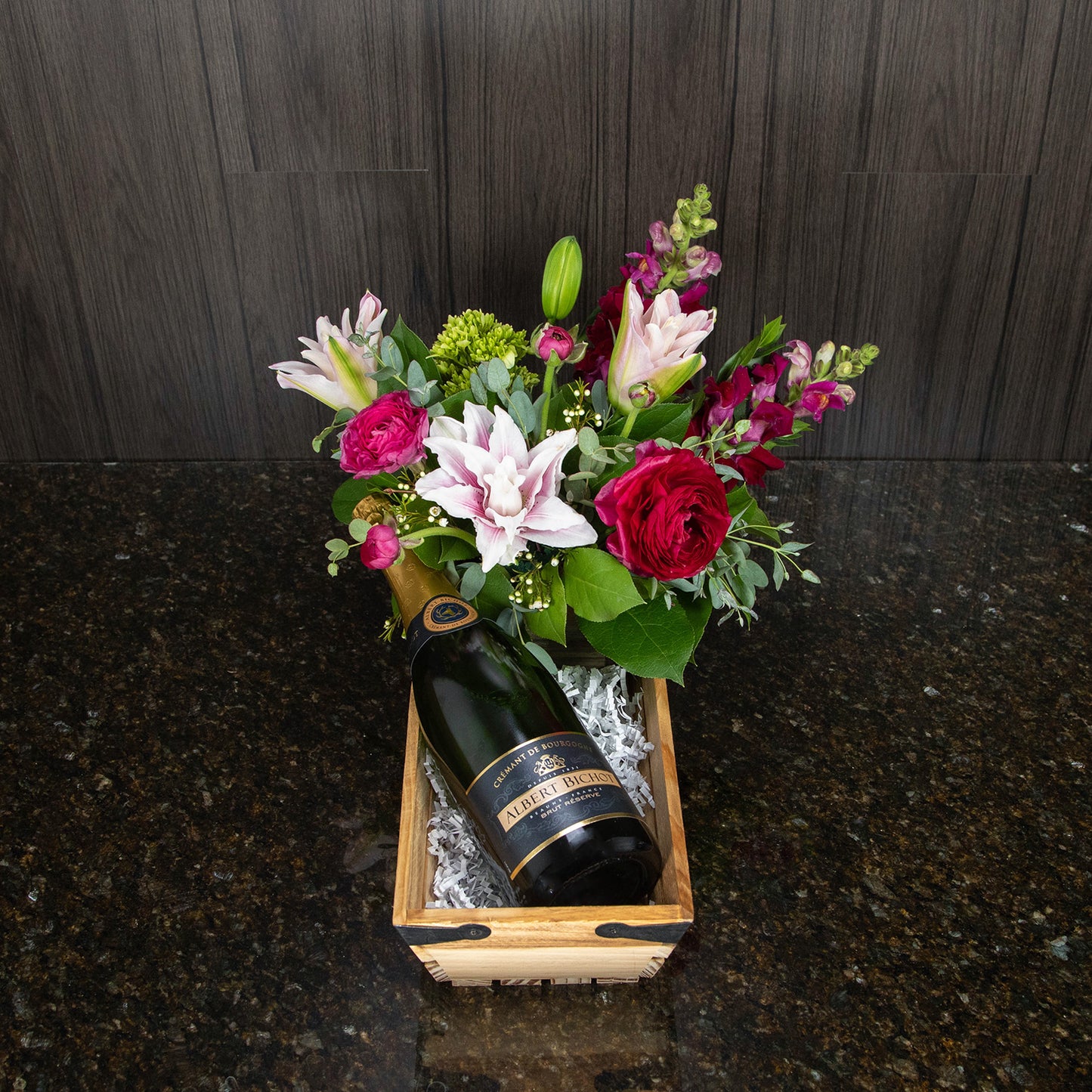 a wooden basket that contains a flower arrangement and a bottle of sparkling wine
